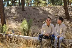 Family photo shoot. Two young brothers wearing matching beige sweaters in a fall setting. They are sitting on a fallen tree trunk behind light foliage, having a fun conversation together.