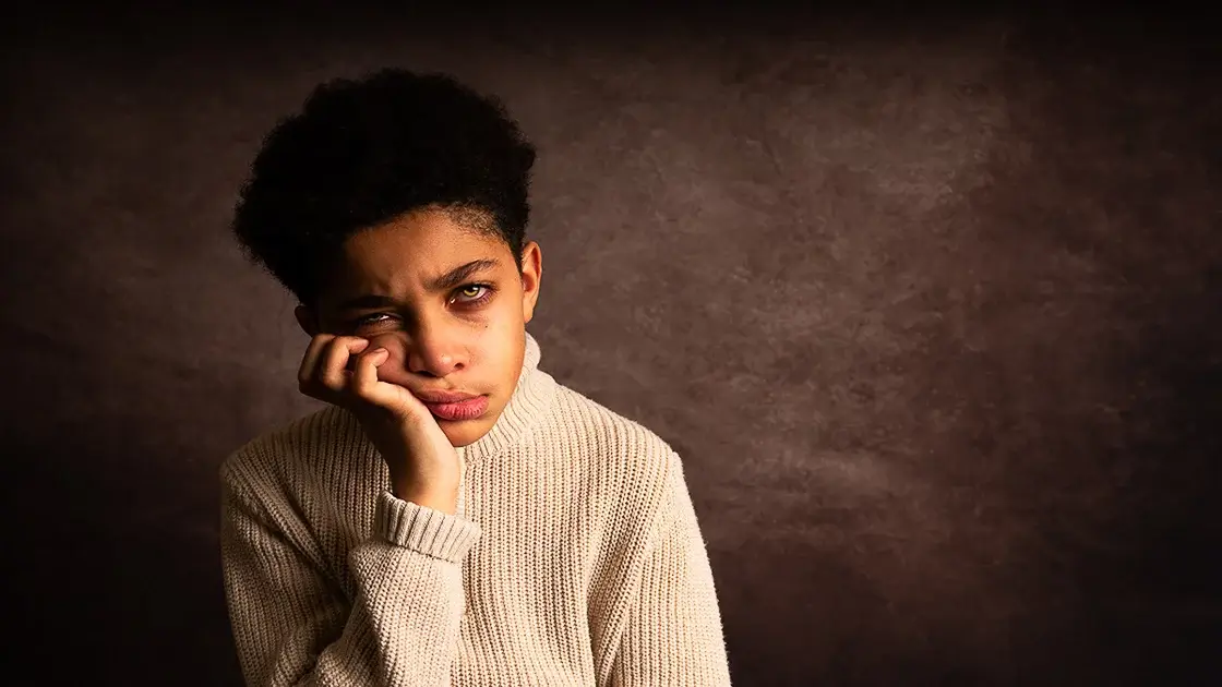 Portraits. A dramatic portrait of a young boy with striking eyes that convey the emotion that he is feeling. He is frowning with his chin resting in his hand. He looks disgruntled or disappointed. He is wearing a beige sweater and is seated to the left side of the image against a brown backdrop.